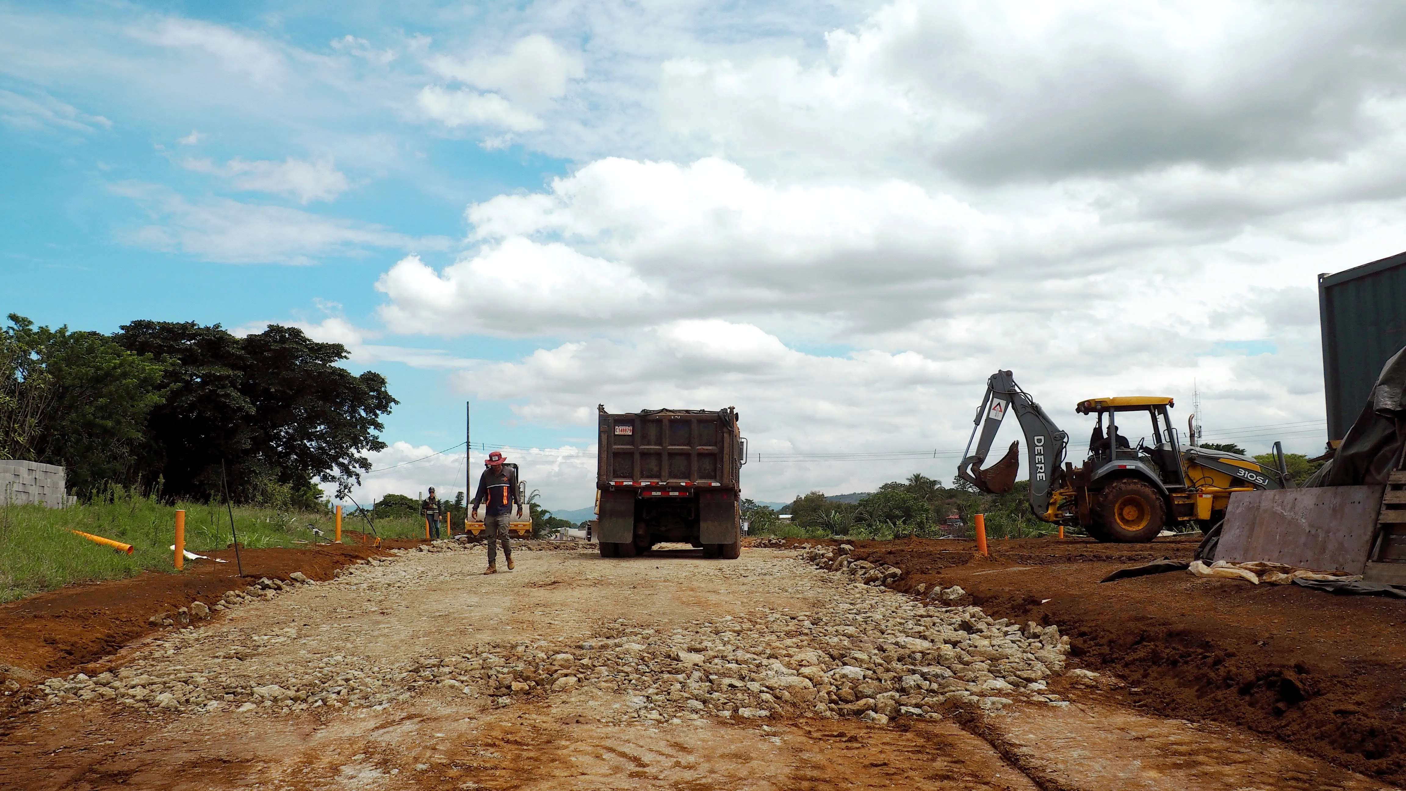 A worker crossing a gravel construction site past a dump truck and excavator under a bright sky
